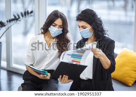Two women checking charts and discussing business trends in the office. Females colleagues wearing medical masks sit on the couch at a working place. Coronavirus prevention. Colaboration concept