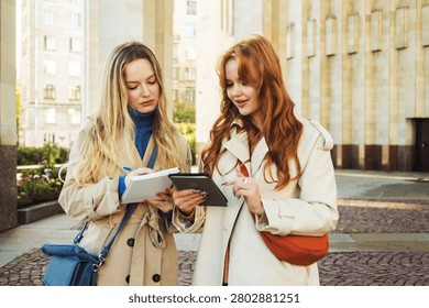 Two women compare notes or documents while standing outside, appearing focused and friendly.