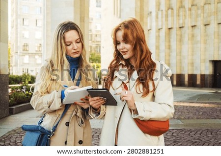 Two women compare notes or documents while standing outside, appearing focused and friendly.