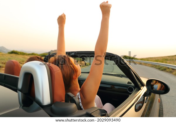 Two women in a convertible car driving and raising\
arms in a mountain road