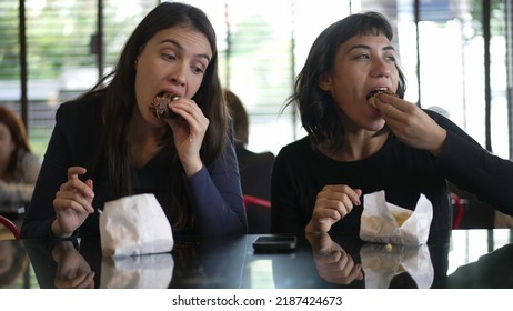 Two Women Eating Burgers For Lunch. Female Girlfriends Taking A Bite Of Hamburgers Together At The Same Time