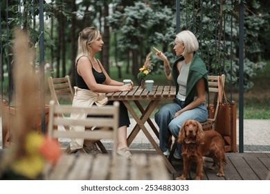 Two women enjoy a conversation at an outdoor cafe. A brown dog sits beside them, creating a relaxed, friendly atmosphere. They are surrounded by nature in a tranquil setting. - Powered by Shutterstock