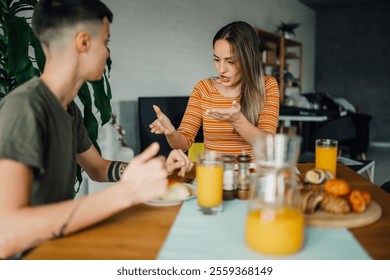 Two women are enjoying breakfast at their dining table, engaging in a lively conversation while savoring pastries and orange juice in the comfort of their modern apartment - Powered by Shutterstock