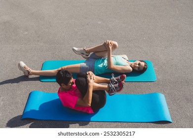 Two women exercising on yoga mats outdoors, performing side plank poses. One woman is smiling and wearing a pink shirt, while the other is in a light blue tank top. Bright and cheerful atmosphere. - Powered by Shutterstock