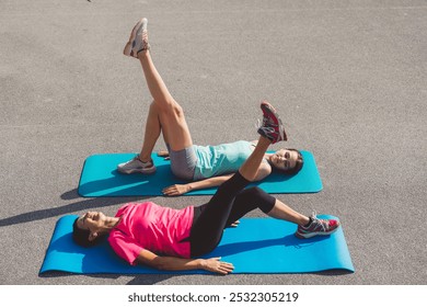 Two women exercising on yoga mats outdoors, performing side plank poses. One woman is smiling and wearing a pink shirt, while the other is in a light blue tank top. Bright and cheerful atmosphere. - Powered by Shutterstock