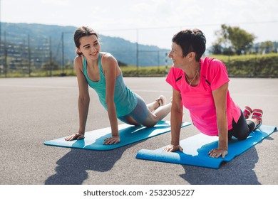 Two women exercising on yoga mats outdoors, smiling and engaging in conversation. They are in a fitness setting with a sunny sky and green hills in the background. - Powered by Shutterstock