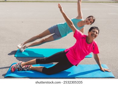 Two women exercising on yoga mats outdoors, performing side plank poses. One woman is smiling and wearing a pink shirt, while the other is in a light blue tank top. Bright and cheerful atmosphere. - Powered by Shutterstock