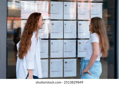 Two Women Exploring at Advertised Properties, Trying to Find House to Buy or Rent on Bulletin Board Outside Building an Urban Setting