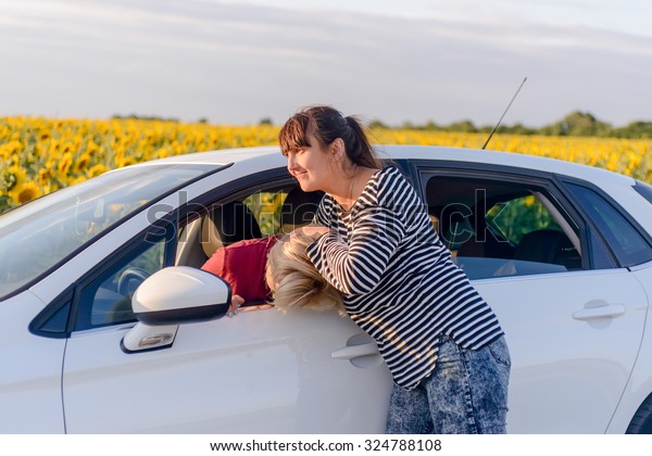 Two women fighting in a car with one\
standing outside in the road gripping the female driver by the hair\
- car parked on a rural road alongside\
sunflowers