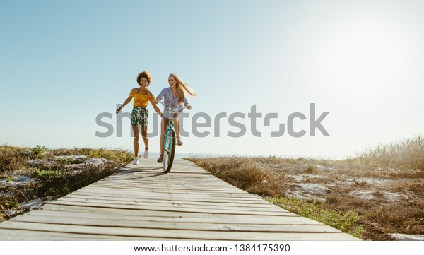 Two women friends with a bicycle on the seaside\
boardwalk. Woman riding the bike with her friends holding from the\
back and running.