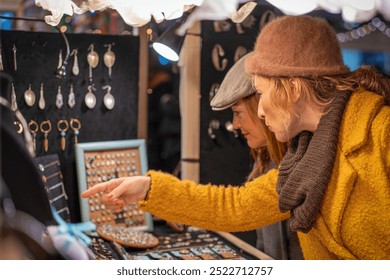 Two women friends shopping for jewelry at a Christmas market. Browsing handmade accessories, wearing winter coats and hats. Festive atmosphere with lights and decorations. - Powered by Shutterstock