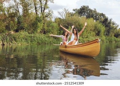 Two women happily raise their arms while sitting in a wooden canoe on a serene river, surrounded by lush greenery on a clear, sunny day. - Powered by Shutterstock