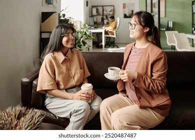 Two women having coffee and conversation in office lounge area, exchanging smiles and ideas while sitting on comfortable chairs in a well-lit office environment - Powered by Shutterstock