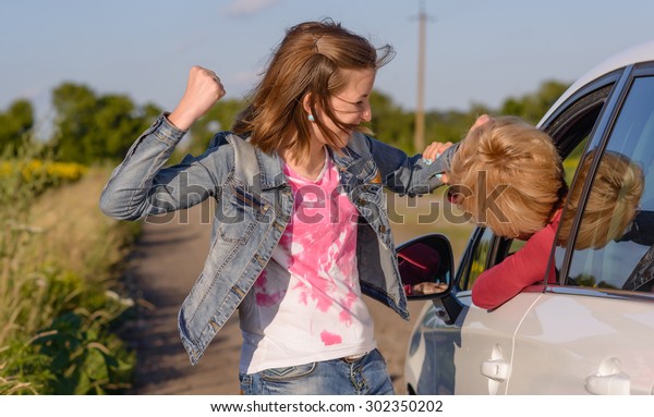 Two women having a fight with a young woman standing\
on the roadside punching the female driver in a car on the head,\
rural road