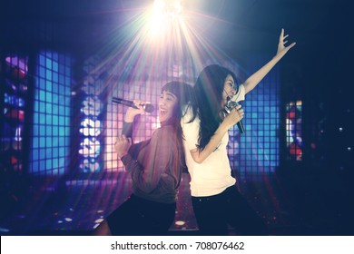 Two Women Having Fun Together In The Night Club While Singing Under A Disco Ball With Bright Rays