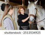 Two women interact with a white horse inside stable, one gently patting horse on nose while the other watches. Wooden stable walls and horse equipment are visible in background