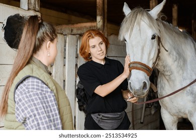 Two women interact with a white horse inside stable, one gently patting horse on nose while the other watches. Wooden stable walls and horse equipment are visible in background - Powered by Shutterstock