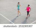 Two women jogging together on a sports court, one in a light blue tank top and shorts, the other in a pink shirt and black leggings. They appear to be enjoying their exercise routine.