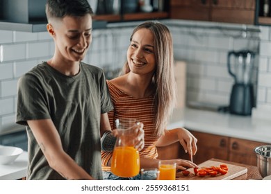 Two women joyfully preparing a healthy breakfast together in a modern kitchen, savoring orange juice and fresh tomatoes while sharing laughter and affection - Powered by Shutterstock