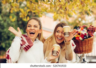 Two Women Laughing With A Blanket On A Bench Enjoying Coffee