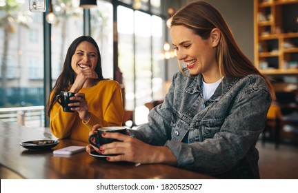 Two Women Laughing Inside A Coffee Shop. Friends Sitting With Coffee Cups On The Table Having Fun.