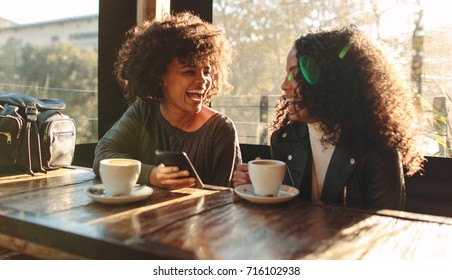 Two Women Laughing Looking At A Mobile Phone Inside A Coffee Shop. Woman Sitting With Coffee Cups On The Table Having Fun.