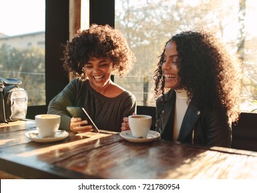 Two Women Laughing Looking At A Mobile Phone Inside A Coffee Shop. Friends Sitting With Coffee Cups On The Table Having Fun.