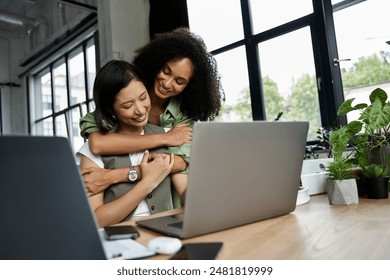 Two women, a lesbian couple, embrace while working on a laptop in a bright office. - Powered by Shutterstock