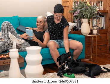 Two women, a lesbian couple, sitting comfortably on their sofa. One is engrossed in reading an ebook while the other lovingly pets their dog. Modern lifestyle and LGBTQ+ companionship concept - Powered by Shutterstock