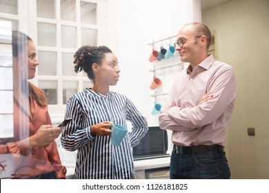 Two women and a man stand and chat together in an office kitchen, one holds a mug the other a phone in a close up