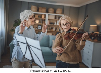 Two women mature female teacher give private violin lesson to her student or friend at home help her to learn to play in room hobby and leisure active senior concept copy space - Powered by Shutterstock