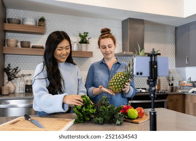 Two women in a modern kitchen preparing fresh vegetables and fruits. Asian woman holds greens, while Caucasian woman holds a pineapple. Diverse people cooking healthy vegetables and fruits in kitchen. - Powered by Shutterstock