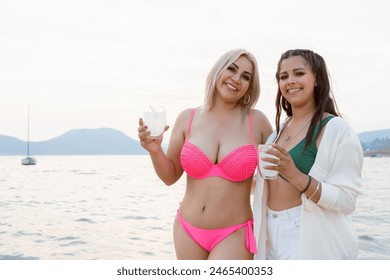 Two women, one in a pink bikini and the other in a green bikini with a white shirt, smiling and holding drinks by the lakeside with mountains and sailboats in the background. - Powered by Shutterstock