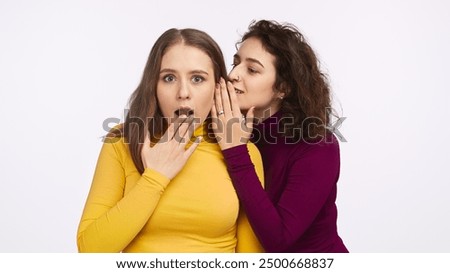 Two women, one in a yellow shirt and the other in a purple shirt, sharing a secret. The woman in yellow shows a surprised expression.
