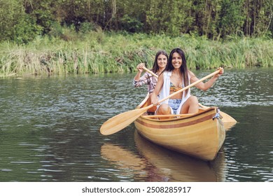 Two women are paddling a wooden canoe on a serene river near green foliage, enjoying an outdoor adventure and bonding time in nature. The canoe glides smoothly on the water. - Powered by Shutterstock