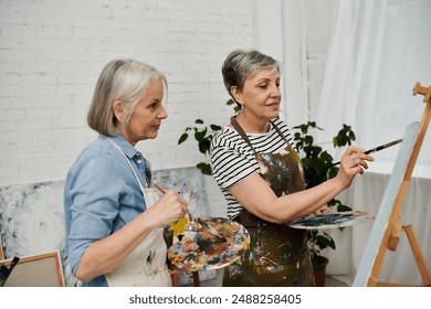 Two women in paint-splattered aprons focus on art in a studio. One paints on a canvas, the other observes and holds a palette - Powered by Shutterstock