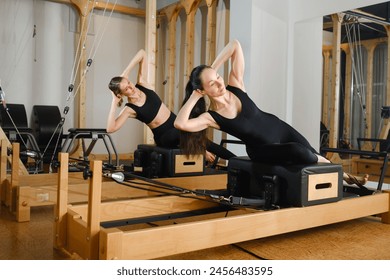 Two women are practicing physical fitness exercises on a Pilates machine in a room with hardwood flooring. Their movements showcase a beautiful balance of art and exercise - Powered by Shutterstock