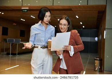 Two women in professional attire converse and review documents in a modern office lobby. - Powered by Shutterstock