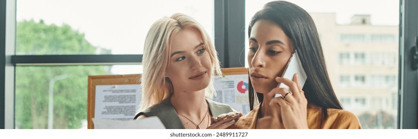 Two women in professional attire work on a project in a modern agency. One talks on phone while other looks focused, lesbian couple - Powered by Shutterstock