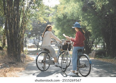 Two women are riding bicycles on a shaded, tree-lined path. They are stopped and facing each other, with one of them holding a map, possibly discussing directions. - Powered by Shutterstock