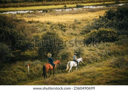 Two women riding a brown and white horse along a trail through a swampy oceanside landscape with small ponds and a river 
