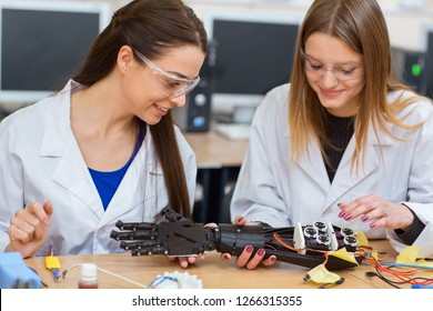 Two women scientists examining mechanical hand in laboratory. Female students, wearing in uniform, studying structure of prototype of mechanical plastic hand robot that can using to prosthetic people. - Powered by Shutterstock