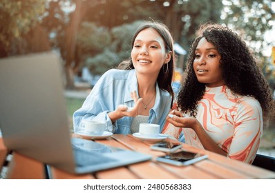 Two women are seated at a table in a cafe setting. One woman is looking at the laptop and speaking, while the other woman listens intently. Both women have coffee cups in front of them. - Powered by Shutterstock