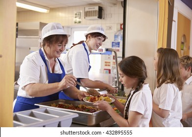 Two women serving food to a girl in a school cafeteria queue