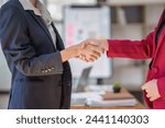 Two women shaking hands in a business setting. The woman on the left is wearing a black jacket and the woman on the right is wearing a red jacket