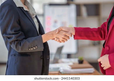 Two women shaking hands in a business setting. The woman on the left is wearing a black jacket and the woman on the right is wearing a red jacket - Powered by Shutterstock