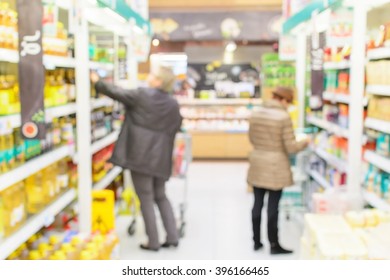 Two Women Are Shopping In A Supermarket In Blurry Effect Background. Customer Choosing And Reading Product Label While Shopping. Concept Of Mass Consumerism During A Period Of World Economic Crisis.