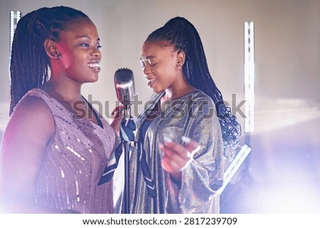 Two women sing into a shared microphone on a lit stage during an evening concert performance as they wear shiny sequined dresses and express their musical talent through powerful vocals