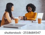 Two women sit across from each other at a table with coffee, engaging in a positive business conversation. Laptops and documents are present, indicating a professional environment.