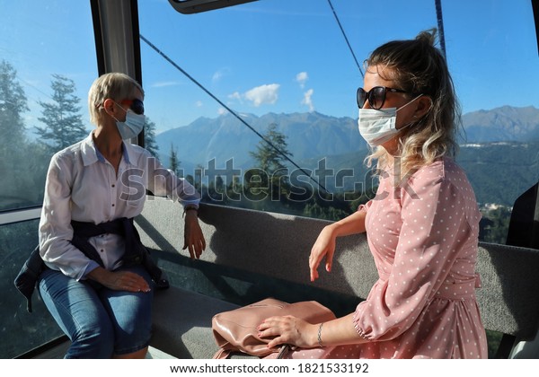 Two women sit in glassy cabin cable car against the\
backdrop of mountains. They are wearing medical face masks and at\
distance. Social distance after quarantine during travel in\
selective focus